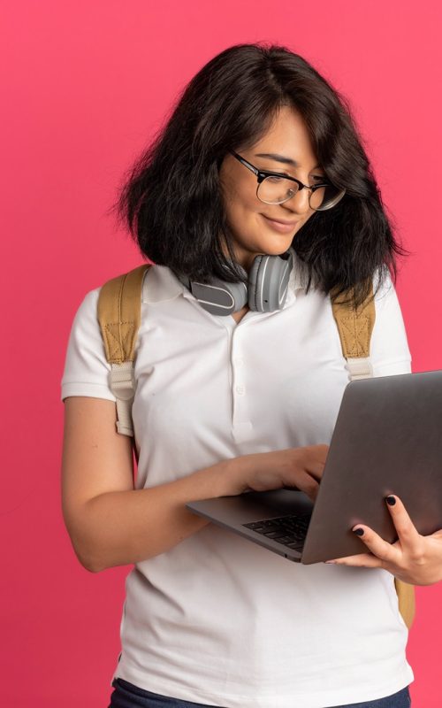 young-pleased-pretty-caucasian-schoolgirl-with-headphones-neck-wearing-glasses-back-bag-holds-looks-laptop-pink-with-copy-space (1)